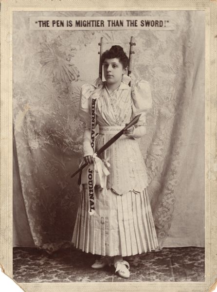 Full-length portrait of a woman posing in a costume made out of newspapers from the "Minneapolis Journal." She is holding an oversized ink pen and has what appears to be telegraph or electrical wires with poles behind her head. A purse hangs from her belt and her shoes are tied with bows. She is standing in front of a figured drape backdrop. Above her is a banner that reads, "The Pen Is Mightier Than The Sword!."