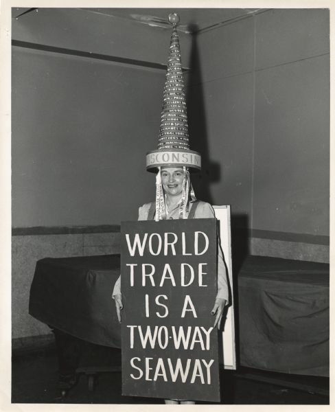 A woman dressed in a costume that would appear to represent businesses and products of Wisconsin. Her very tall hat has a world globe on the top and the word "Wisconsin" around the brim. Words describing the products of Wisconsin wind around the hat. Ribbons dangling on both sides of her face have the names of Wisconsin companies, Parker Pen Co., Pfister & Vogel, Caterpillar Tractor, Allis Chalmers, an Iron Works Company, etc. She is wearing a sandwich board sign that reads, "World Trade Ia A Two-Way Seaway." Behind her under a cover is a grand piano.