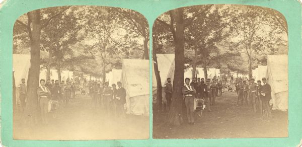 Quite possibly this is the "General view in the camp" listed in the "Centennial Views of the City of Madison, July 4th, 1876. Views of Chicago Light Guard" section of Dahl's 1877 "Catalogue of Stereoscopic Views." The men are encamped at Camp Randall or on the shores of Lake Monona. The group of men are posing between two rows of tents, some of which have American Flags. A young man with a drum leans against a tree in the foreground.