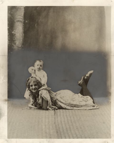 Actor John Gilbert as a baby with his mother. She is lying on the floor on her stomach, resting on her elbows with her head up. Her knees are bent and her ankles are crossed. He is standing behind her head.