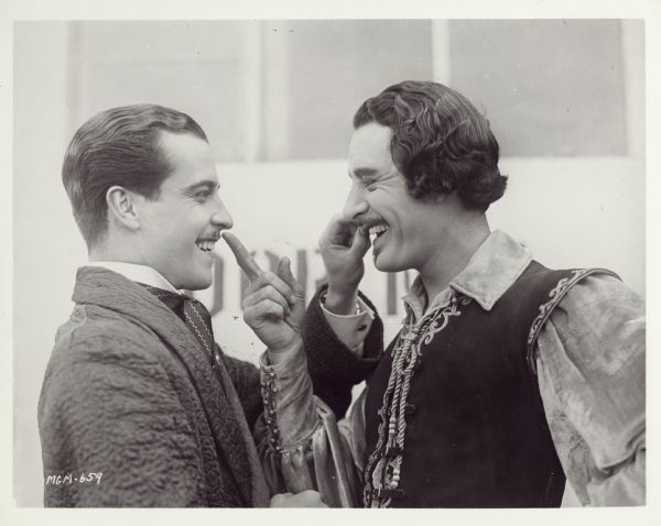 Actors John Gilbert and Ramon Novarro compare their moustaches. Gilbert is in costume.