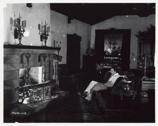 Actor John Gilbert sitting on a couch in the living room of his Beverly Hills mansion. He is staring at the fireplace which has a light fixture inside it.