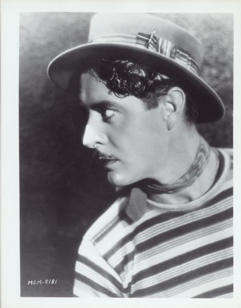 Publicity still for the film "The Show." John Gilbert is seen in profile — he is wearing a hat, striped shirt, and a scarf around his neck.