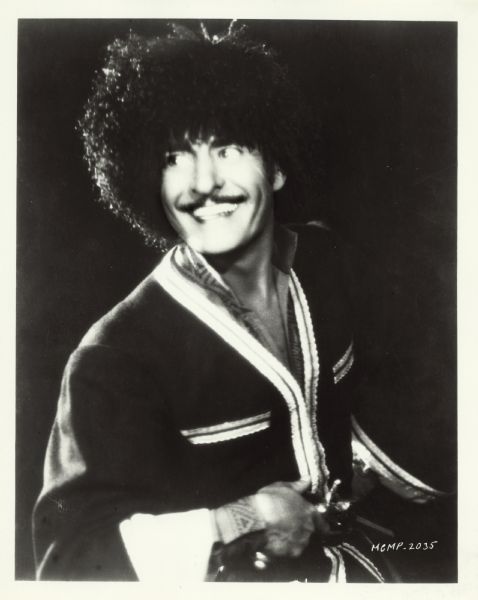 Publicity still of actor John Gilbert in "The Cossacks." He is looking towards the left and smiling, and is wearing a big bushy hat and holding a gun at his waist.