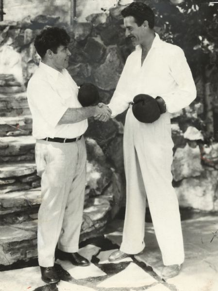 Actor John Gilbert and boxer/author Jim Tully shake hands. Each is smiling and wearing a boxing glove on one hand. This publicity still may be a reference to an actual fight between Gilbert and Tully in the Brown Derby restaurant in 1930.