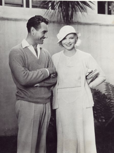 Three-quarter length portrais of actor John Gilbert and his fiancée Virginia Bruce. Both are smiling — he is looking at her and she is looking at the camera.  They are holding hands.