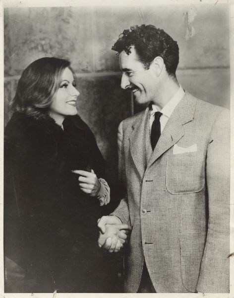 Actor John Gilbert and Greta Garbo shake hands and smile at one another their first day on the set of the movie "Queen Christina." She is wearing a fur coat and he's in a three-piece suit.