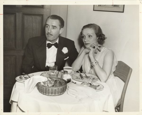 Actor John Gilbert sits next to Marlene Dietrich at a party thrown by Clifton Webb. They are both in formal dress. There are empty dinner plates and a basket of rolls on the table.