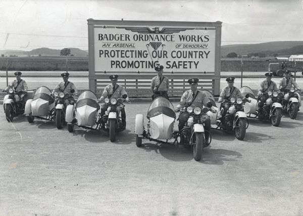 Motorcycle police at the Badger Ordnance plant in Baraboo, Wisconsin, between 1942 and 1945. These men patrolled the fence line of the plant. One of the guards is Lyle Lilly (1916-1911). The sign reads, "Badger Ordnance Works, An Arsenal of Democracy, Protecting Our Country, Promoting Safety." There is a tractor in the right background.