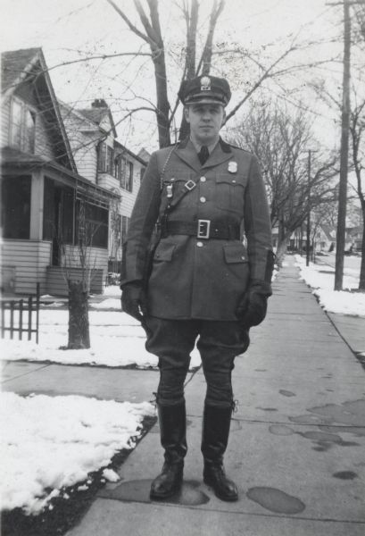 Lyle Lilly (1916-2011) stands in front of his home at 838 Woodrow Street in Madison, Wisconsin. He is wearing his motorcyle police uniform that he wore while working at the Badger Ordnance Plant in Baraboo, Wisconsin.  Motorcycle police patrolled the fence line at the plant.  Snow is on the ground.
