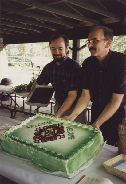 Brian Bigler (right) and Ken Scott (left) on July 22, 1995 at the reception for their Holy Union Ceremony, which was held at Brigham Park, Blue Mounds, Wisconsin.  The couple are posed in a picnic pavilion with their green Holy Union cake.  The decoration on the cake was taken from their altar cloth.  Ken is also holding a pan of brownies.