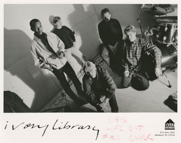 Publicity photograph of five band members from Ivory Library, a Madison, Wisconsin rock band. From left to right are Derrick McBride, Dave Batson (standing against the wall), Jeff Jagielo (seated in front), Jim Luepton, and Peter Bruhn (leaning against the bass drum). The band was on the Boat Records & Tapes label.