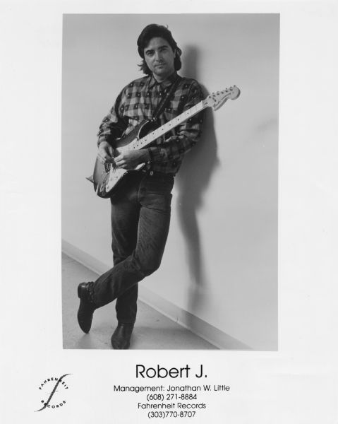 Publicity photograph of musician Robert J. of Madison, Wisconsin. He is holding a Fender Stratocaster electric guitar while leaning against a wall with his legs crossed.<p>Fahrenheit Records is mentioned on the photograph.