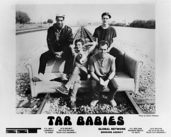 Publicity photograph of the Tar Babies, a Madison, Wisconsin rock band. From left to right are Tony Jarvis, Bucky Pope, Robin Davies, and Dan Bitney. They are sitting on a sofa placed on railroad tracks. The band was on the SST Records label.