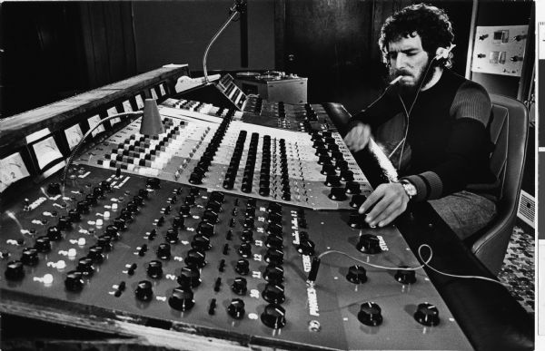 Musician Ben Sidran sitting in front of a console in a music studio. He is wearing earphones and smoking a cigarette. Ben has a moustache and beard.