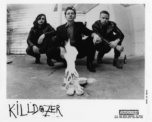 Publicity photograph of the three band members of Killdozer, a Madison, Wisconsin rock band. From left to right are Bill Hobson, Michael Gerald (holding cigar), and Paul Zagoras. All three are squatting in front of a stuffed animal, possibly a teddy bear, that's on fire. A Leinenkugel beer bottle is at the right.<p>Killdozer is on the record label Touch and Go Records.


