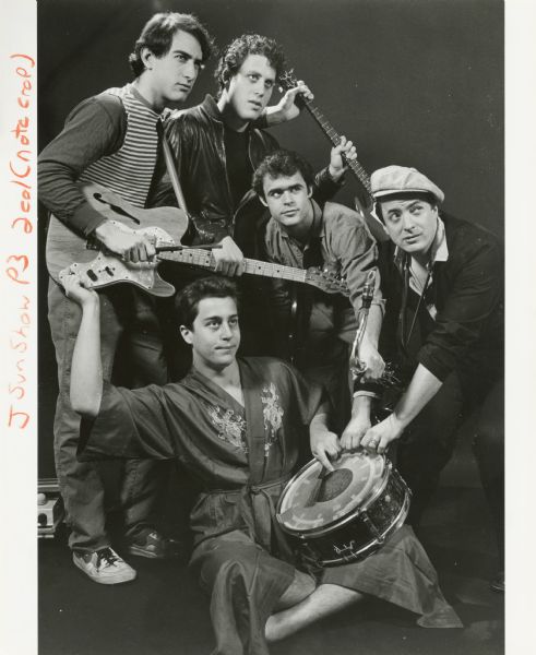 Publicity photograph of the five band members of Swamp Thing, a Madison, Wisconsin rock group. Clockwise from top are Jonathan Zarov, Bob Appel, Mike Kashou, David Miller, and Steve Bear (seated). They are holding two electric guitars, a saxophone, and snare drum. Steve is wearing a silk bathrobe with an embroidered dragon design.

