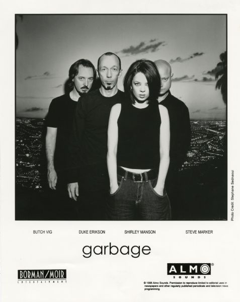 Publicity photograph of band members from Garbage, a Madison, Wisconsin rock band. From left to right are Butch Vig, Duke Erikson, Shirley Manson, and Steve Marker. They appear to be standing in front of a photograph of a city from a high elevation.<p>Mentioned on the photograph is Borman/Moir Entertainment and Almo Sounds.