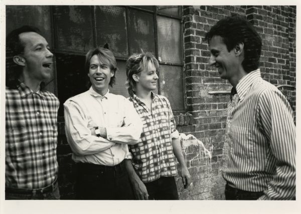 Publicity photograph of the band members from Fire Town, a Madison, Wisconsin rock band. From left to right are Doug Erikson, Phil Davis, Butch Vig, and Tom LaVarda. They are standing outside of a brick building, possibly abandoned, in front of a window with cracked and broken glass.