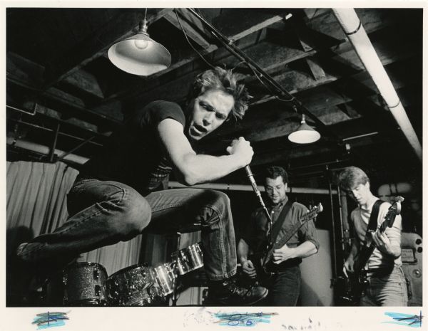 The rock band Appliances-SFB playing in guitarist William Siebecker's basement. Singer Tom Laskin is jumping in the air while holding a microphone. Band members William Siebecker (center) and Ed Feeny (right) are to Tom's right playing electric guitars. A drum kit is in the background.