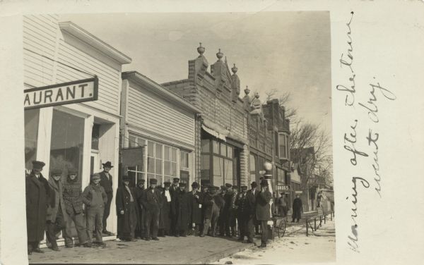 A group of men standing on the sidewalk outside a local business. The sign on the far left of the image is for a restaurant, and the other buildings further down the street are for a real estate company, a grocer and a drugstore. The caption on the image reads "Morning after the town went dry."