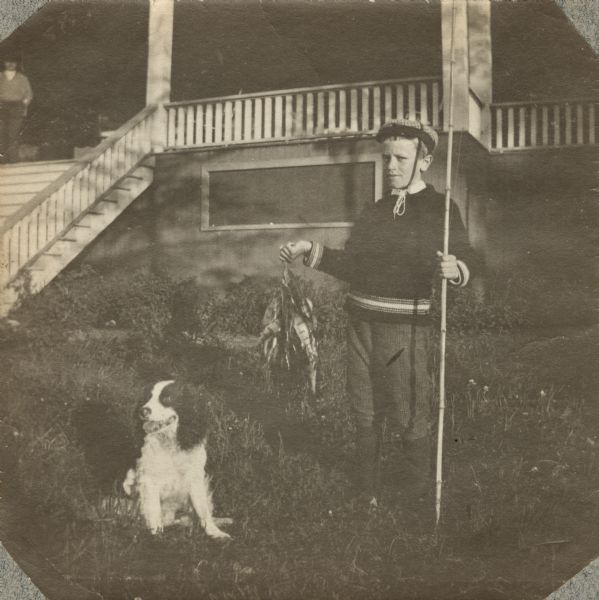 A young Aldo Leopold posing proudly with his bamboo fishing pole, stringer of perch and a dog. The family was on vacation in the Cheneaux Islands, near Mackinac. A large porch and stairs are behind him, possibly the Les Cheneaux Club clubhouse. Another figure can be seen in the upper left corner.