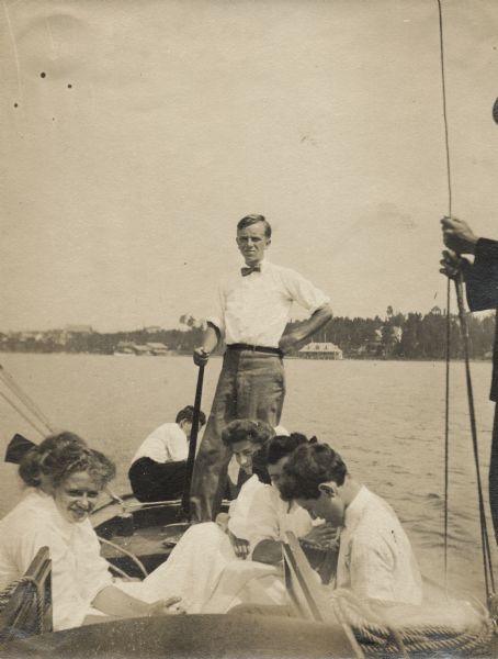 Aldo Leopold standing in a boat with a group of young people. He is wearing pants, a shirt and a bow tie. Buildings can be seen on the shoreline in the background.