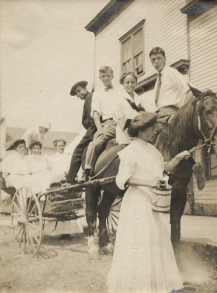 A young Aldo Leopold sitting on a horse with several friends. Handwritten next to the snapshot: "a heavy load." A woman in a dress is holding the horse's lead rope and three women are sitting in the buggy harnessed behind. Another person may be standing in the back of the buggy. There is a building in the background.