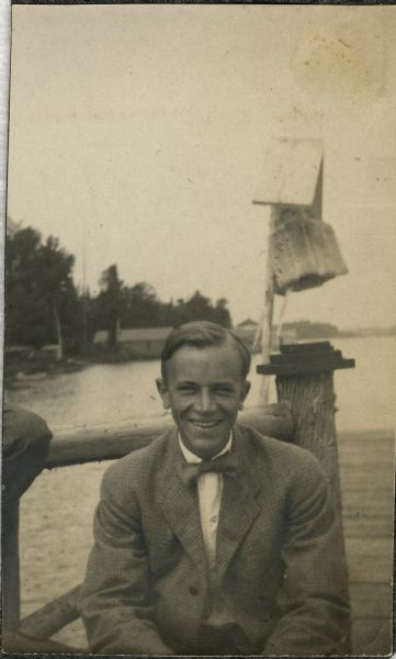 A young Aldo Leopold is posing smiling, wearing a jacket and bow tie while sitting on a pier. He is vacationing in the Cheneaux Islands. A shoreline with buildings is behind him.