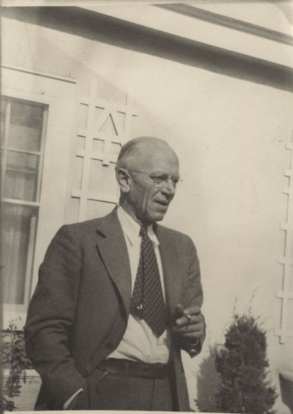 Three-quarter length portrait of Aldo Leopold taken outdoors by his wife. He is wearing a suit and tie. A window, shrubs and a window box are behind him.