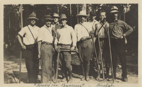 A group of men dressed for work, with implements in hand. Most of them are wearing hats and boots. One man is smoking a pipe. A forest is behind them. Handwritten at bottom: "'Ready for Business' and Pinchot." Note on back, "Aldo 2nd fr. R."
