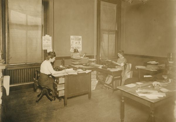 Two women seated at desks with typewriters in an office. A calendar on the wall indicates it is June 1912. Another poster advertises speakers in support of women's suffrage and a suffrage automobile. There are bundles of papers on tables and a Waukesha water cooler at right.