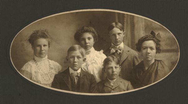 Quarter-length oval studio group portrait of six young people, three boys and three girls. The girls are wearing fancy dresses and the boys are wearing suits. Their names were written in pencil on the wooden back piece of the frame. Some names are not legible. Left to right: Inga E. Karlslyst, Burns Sa__, Ada Gilbertsen or Gilbertson, Fred _abjornsen, Franklyn Iversen or Iverson, Anna J_w_zy Tealf_?