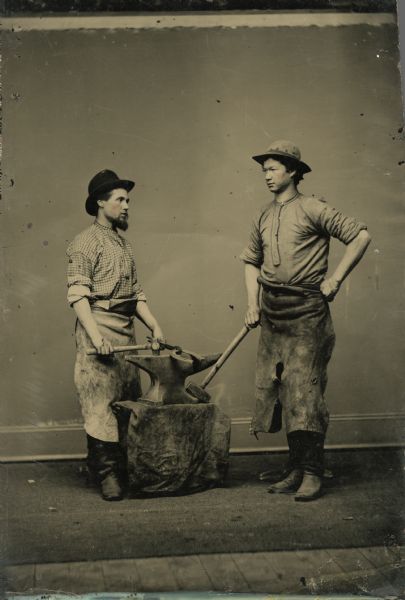 Ferrotype/tintype of a studio portrait of Blacksmith Wanamaker and an unidentified man standing on either side of an anvil. The man on the left is holding a horseshoe with forge tool tongs in his left hand and a hammer in his right. The other man is resting the head of a sledge hammer on the base of the anvil. The anvil support is covered with a dirty cloth. The men are wearing work clothes, leather aprons, boots and hats.