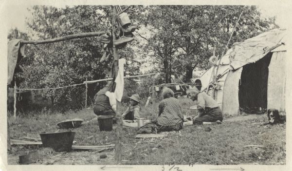 St. Croix Ojibwa Chief Ma-Ko-day (Chief Peter Bearheart) and his wife, grandson, and another unidentified person in front of their birch wigwam located in the Rice Lake Encampment. A dog can be seen on the right and buckets and tubs on the left. Shoes, clothes and various implements are hanging on a pole. There are trees in the background.