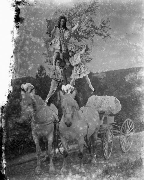 Composite photograph of one man and two women standing on top of two horses pulling a buggy. The three figures are glued onto the photograph containing the horses. The man is wearing a suit, and the women are wearing costume dresses and holding umbrellas. The horses have plumes glued to the tops of their heads. The performers are identified as the Hocums. They performed chiefly with the Gollmar Bros. Circus. The Gollmar Bros. started their own circus in 1891 and operated until 1916, spending the winters in Baraboo, Wisconsin. After 1916 the Gollmar name was leased to other circuses, and last used in 1926.