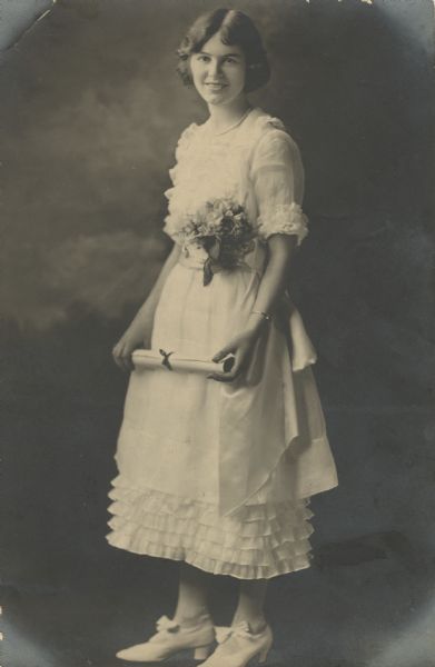 Full-length studio graduation portrait of Genevieve Milward (later McVicar), (1902-1987) of Madison, Wisconsin, standing in front of a painted backdrop. She is holding her diploma and has a bouquet of flowers tucked into the waistband of her light-colored graduation dress. Her shoes are light-colored with bows and low heels. She is wearing a string of pearls around her neck. 

She graduated from Madison High School in June 1921.

The dress was made by Benevieve's aunt Elizabeth (Meibohn) Milward (1879-1966).

The dress is in the collections of the Wisconsin Historical Museum (2012.139.1a-b).