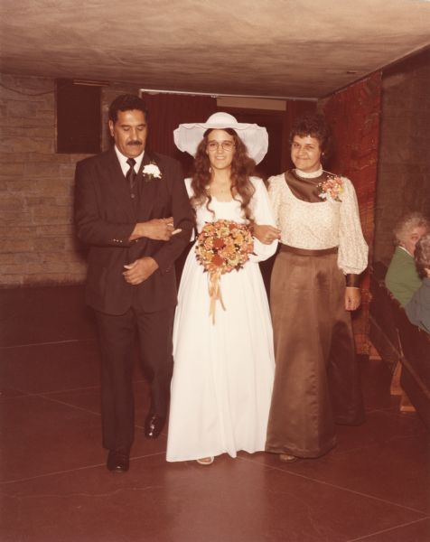 Full-length group portrait of a bride and her mother and father walking towards the center aisle of the First Unitarian Society Meeting House as the ceremony begins.