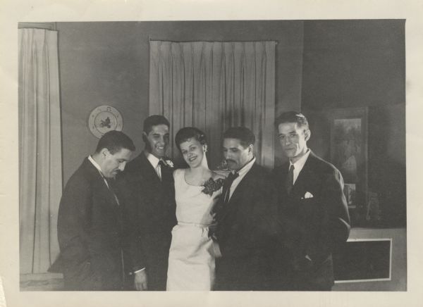 Three-quarter-length group portrait of a bride and groom with three of his brothers. She is wearing a street-length dress of ivory florentine, black shell hat and a corsage of red roses. The men are wearing dark suits.