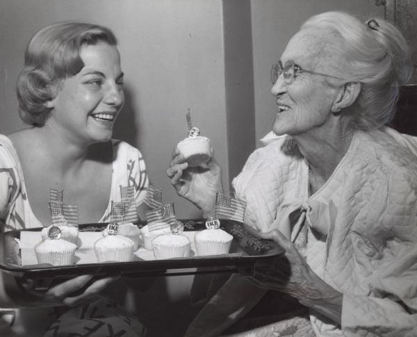A young woman and an elderly woman are smiling at each other over a tray filled with cupcakes decorated with "IKE in '56" campaign buttons and tiny American flags. The young woman is wearing a dress, and the elderly woman is wearing a quilted bed jacket tied with a bow.