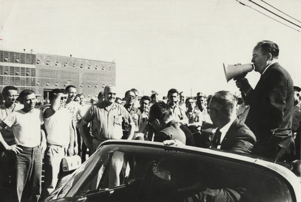 Hubert H. Humphrey speaking to factory workers through a megaphone from the back of a convertible.