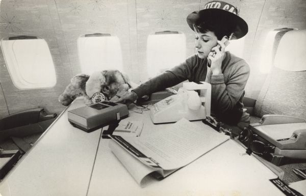 A woman working on a Goldwater campaign airplane. She is wearing a Goldwater hat, and a stuffed elephant is sitting on the desk with a Goldwater pin attached to it. Copy attached to photograph reads: "Any one of the four desks aboard the Goldwater campaign plane is the center of busy work-in-the-air, whether it be typing or listening to dictation. This modern flying communications center contains a radio-telephone air to ground, radio transmitter, modern Dictaphone dictating equipment, mimeograph facilities, intercom system, and full secretarial services."