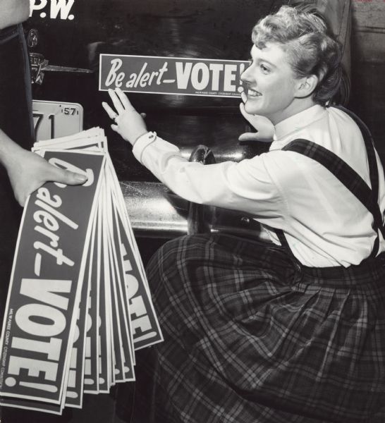 Two U.W. Milwaukee coeds stopped at the city garage to help employees place "Be alert — VOTE!" bumper stickers on city vehicles. Miss Nancy Breske is applying the sticker and Miss Karel Kasten (only her arm is seen) is holding a stack of the stickers. They were provided by the Milwaukee County Citizenship Commission.