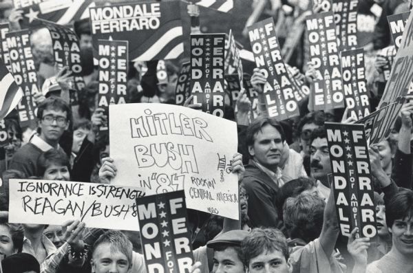 Delegates hold up Mondale/Ferraro signs at the Democratic National Convention. Two are hand-drawn signs, "Hitler/Bush in '84, Rockin' Central America" and "Ignorance is Strength, Reagan/Bush '84."