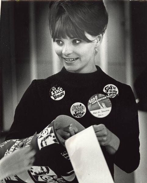 Mary Jo Sylvestor wears campaign buttons supporting Jerry Leonard as the Republican candidate for United States Senate. She is attending the State Republican Convention at the Dane County Coliseum, and is from Madison. On the three campaign buttons is the text, "Even I Like Leonard," "Junkies for Jerry," "Sock it to me, Jerry." On her identification button it says, "G.O.P. Convention, Mary Jo Sylvester, Dane." Around her arm she has banners or bumper stickers. Jerry Leonard lost the election to Gaylord Nelson.