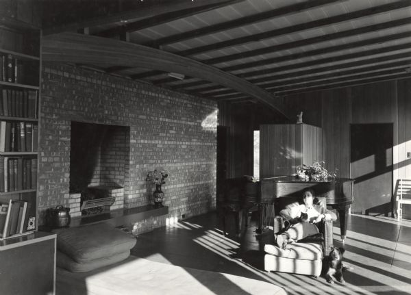 Interior view of the living room of a George Fred Keck designed home. A woman is relaxing in a chair with a cat in her lap, and her dog lies on the floor next to her chair. A grand piano is in the background, and there is a built-in fireplace along the brick wall on the left near a set of bookshelves. Sunlight is coming in from the right casting shadows on the interior.