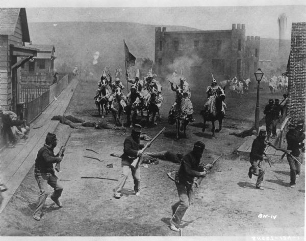 Street scene from the film "The Birth of a Nation" featuring a battle between soldiers on foot and members of the Ku Klux Klan on horseback. The men on horseback are firing guns and one man is carrying a flag. There are bodies on the ground and the men on foot appear to be running away. In the background are men on horses.