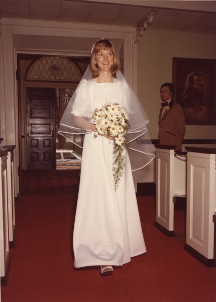 A bride walks down the aisle in the United Church of Christ/Congregational Church in Delavan. An usher stands to the right in the background. A painting of Jesus is behind him. Through the open door, the decorated wedding car can be seen.<p>The bride sewed her wedding dress from a Vogue Couturier Design pattern, #1155, option "C," designed by Belinda Bellville. The pattern was purchased at Northwest Fabrics. The material for the dress was purchased at the Swiss Miss Textile Mart in New Glarus.<p>The wedding dress, veil, and pattern are in the collection of the Wisconsin Historical Society (2012.195.1-2).</p>