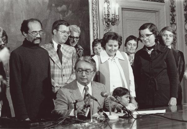 Several men and women watch as Governor Patrick Lucey signs the Equal Rights Amendment. Lloyd A. Barbee (left), Marlin Schneider, two unidentified people, Midge Miller (center), Mary Lou Munts (right), David Clarenbach (far right). Governor Lucey is seated at the desk in the middle.