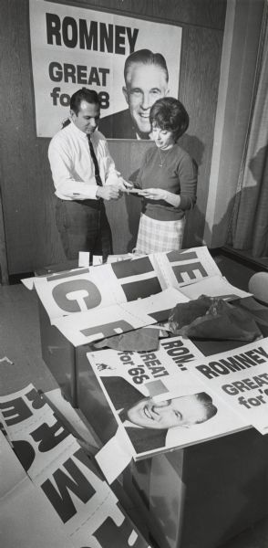 A man and woman get ready for the arrival of Mrs. George W. Romney at the Romney for President headquarters. Several campaign posters and banners are spread around the room — on the wall in the background, on the table and the floor. The slogan "Romney, Great for '68" is printed on several posters.